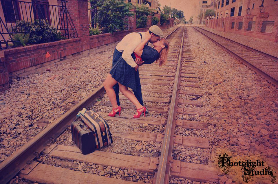 A couple shares a romantic kiss on railroad tracks with a suitcase nearby.