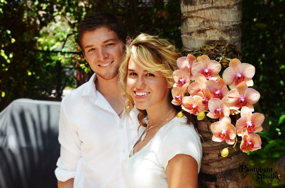 A smiling couple posing outdoors near pink orchids.