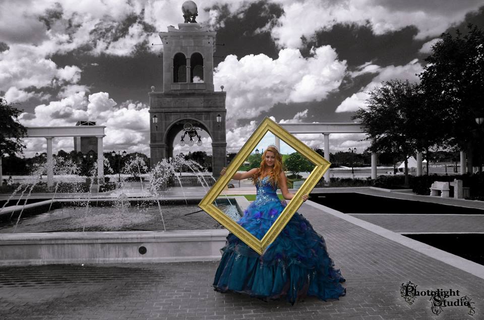Woman in blue dress holding a yellow frame near a fountain and bell tower.