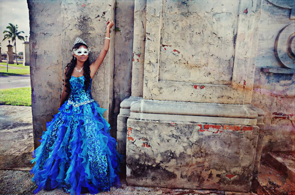 Woman in an elaborate blue gown posing against a rustic wall.