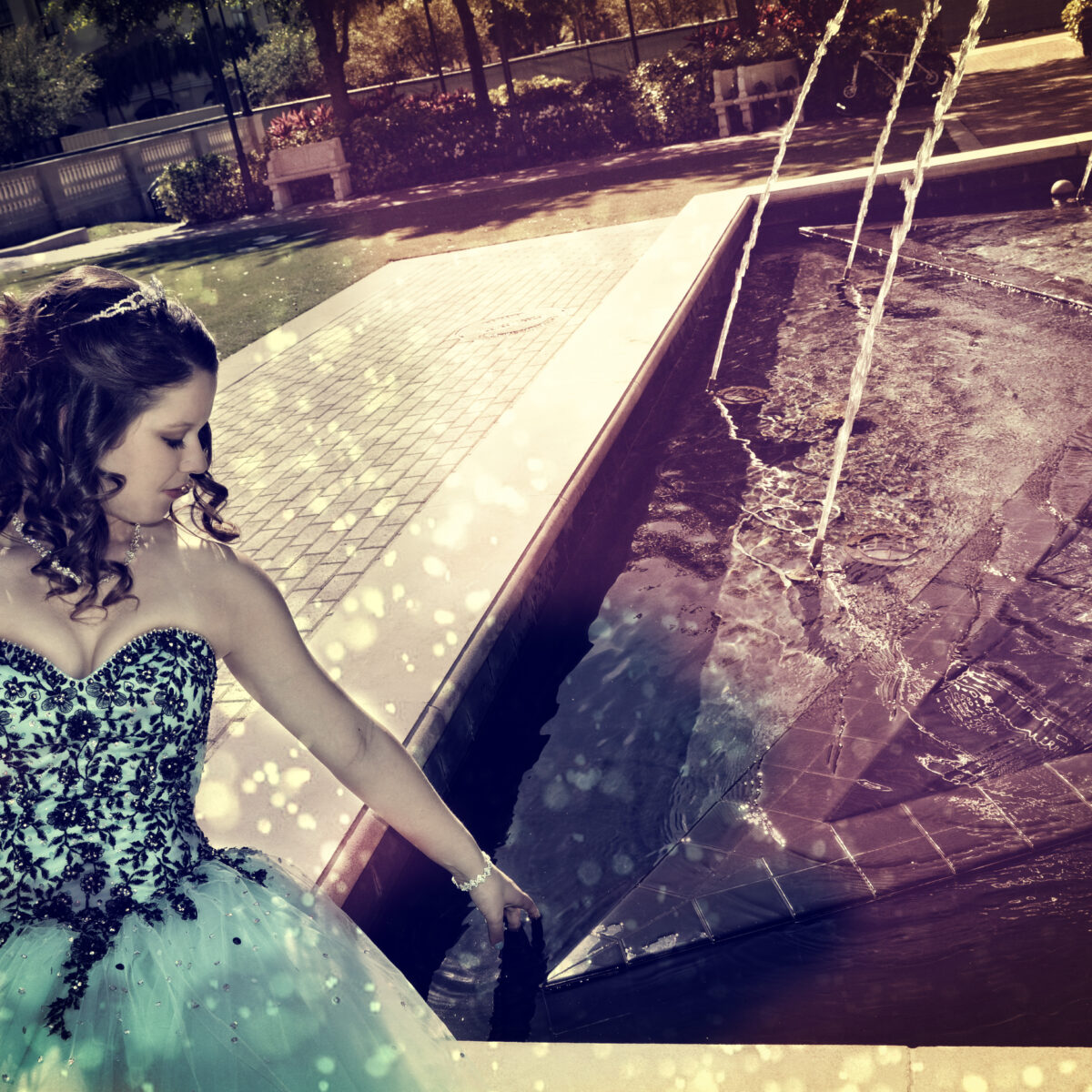 Woman in a turquoise dress near a fountain on a sunny day.