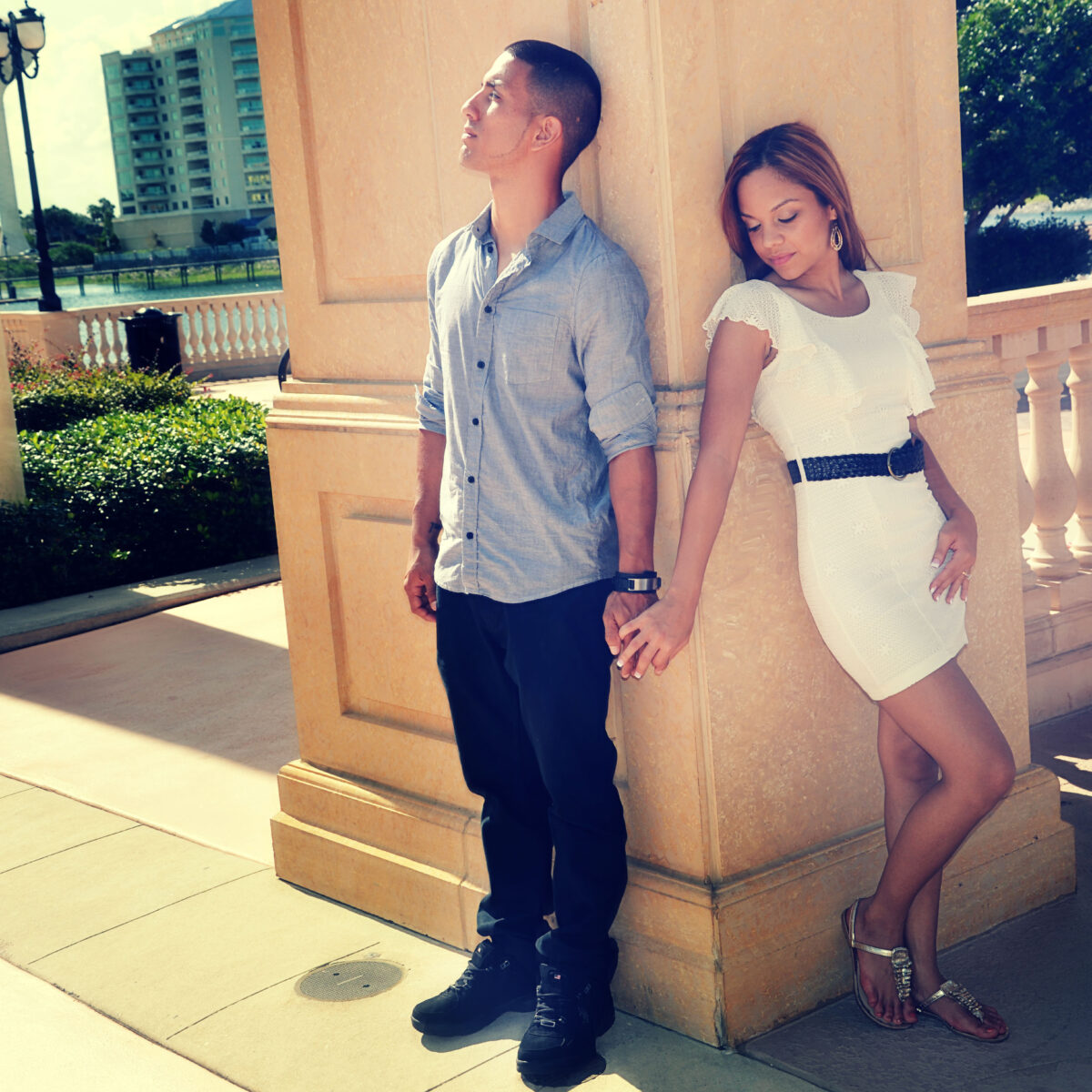 A couple holding hands and leaning against a column outdoors.