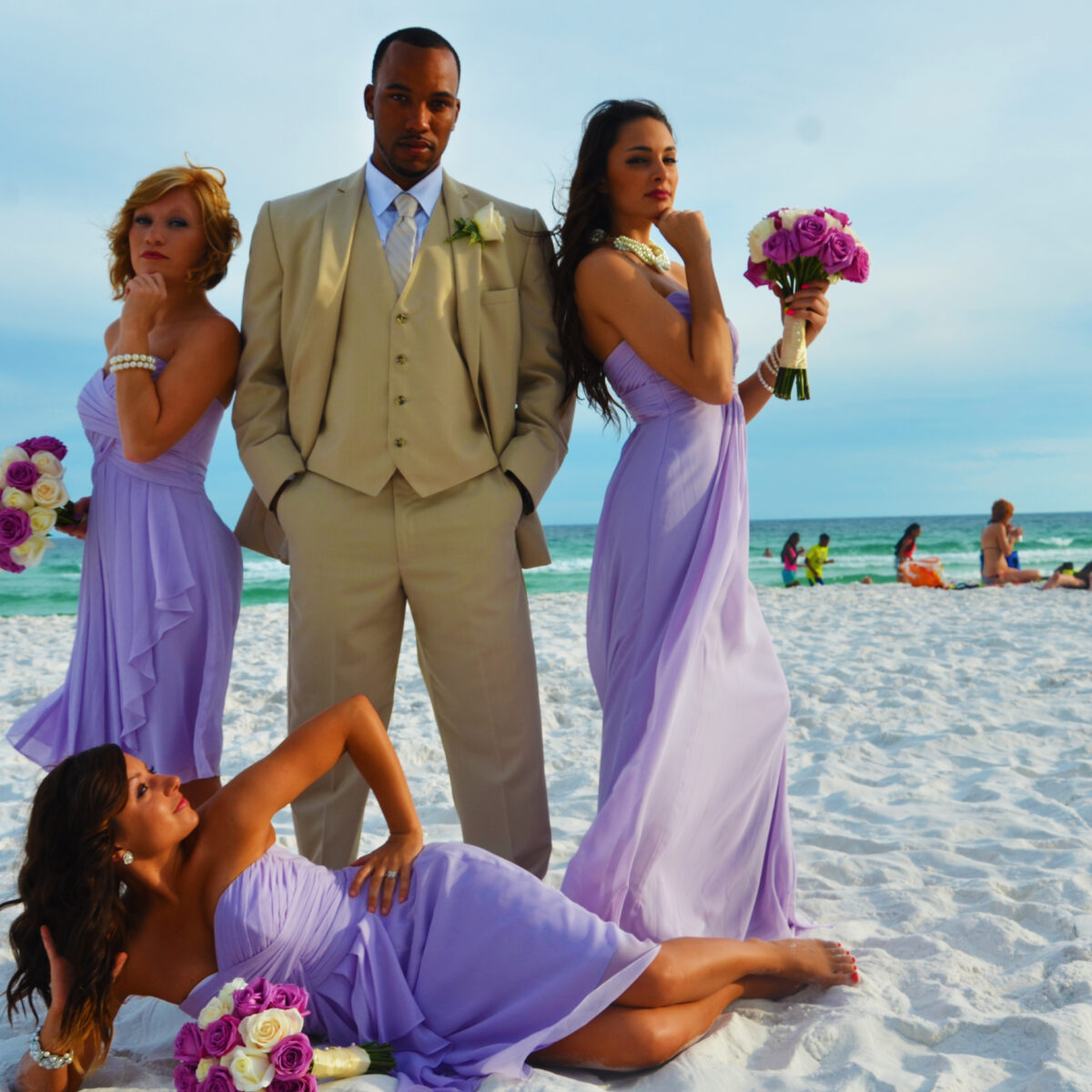 Groom with bridesmaids in lavender dresses posing on beach.