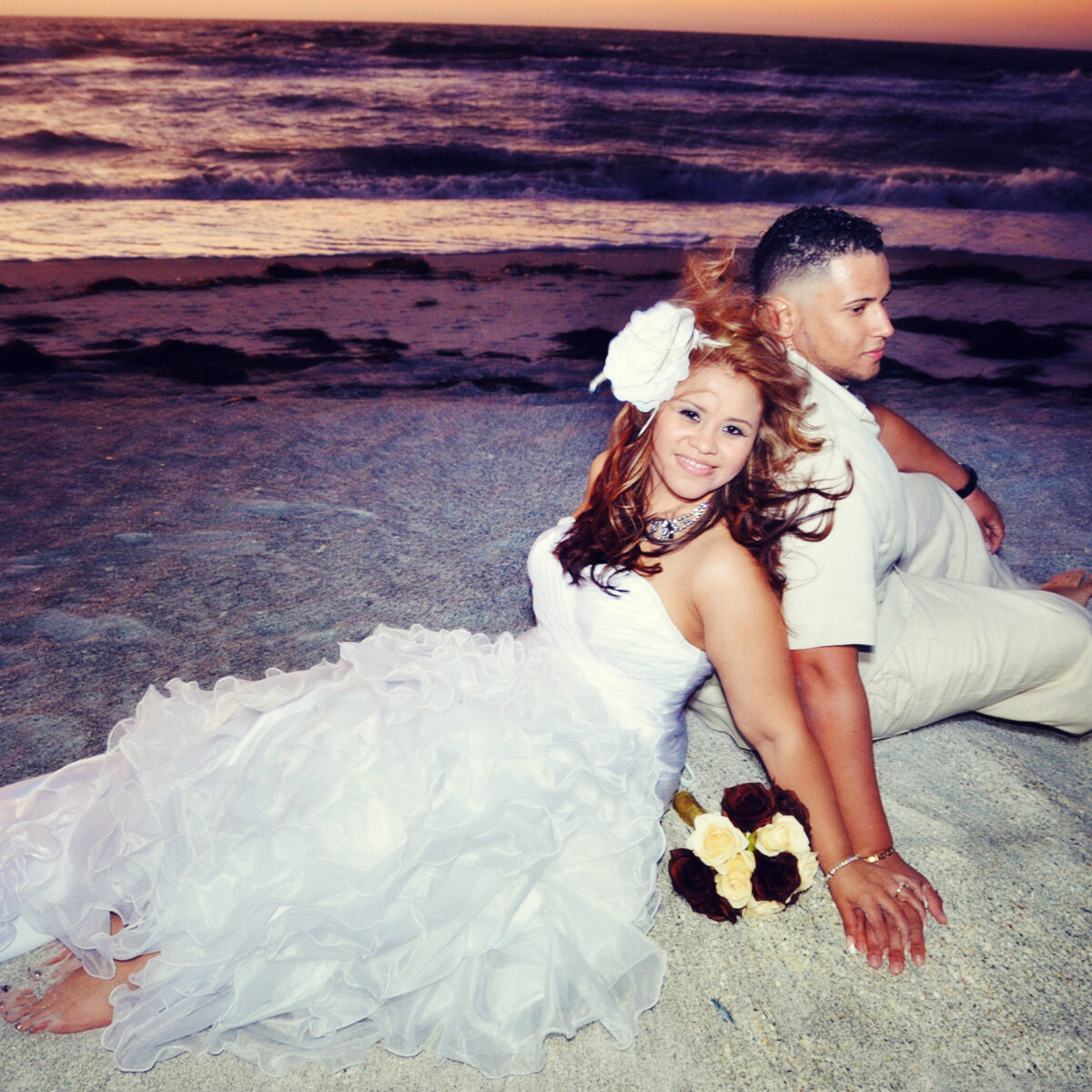 Bride and groom pose on beach at sunset with wedding bouquet.