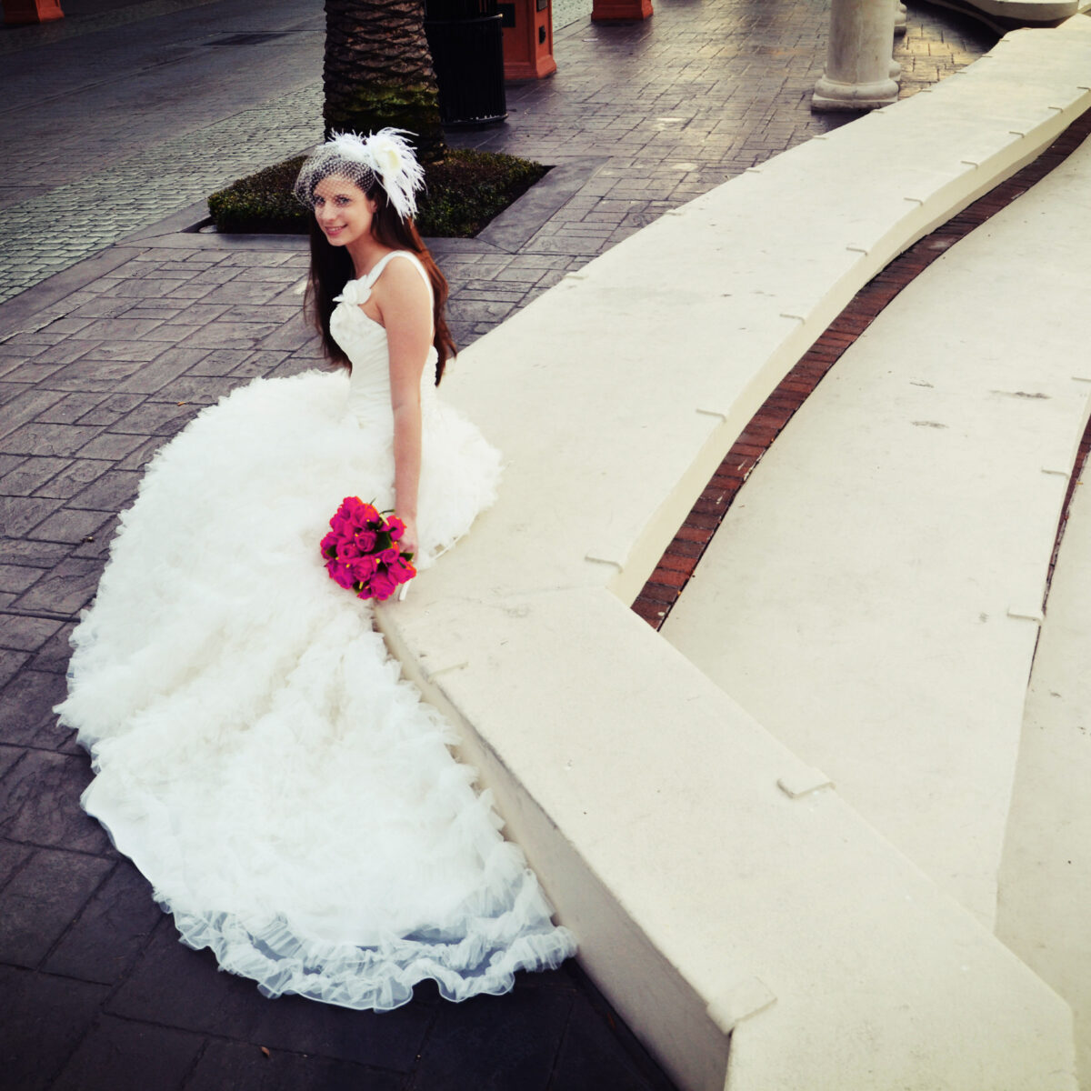 Bride in a white gown holding a pink bouquet, sitting by a curved stone bench.